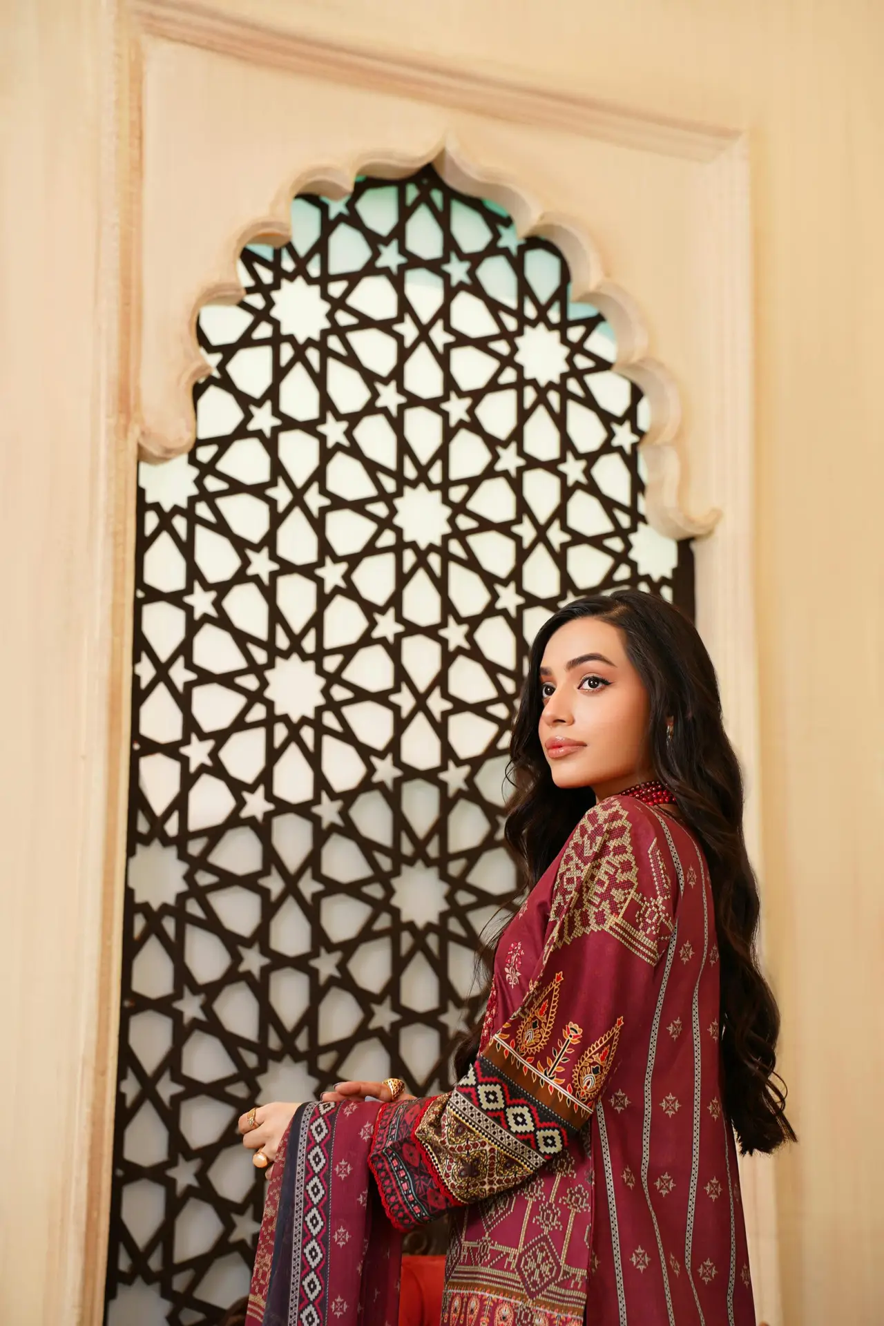 Young woman in a traditional Pakistani dress standing in front of ornate lattice window.